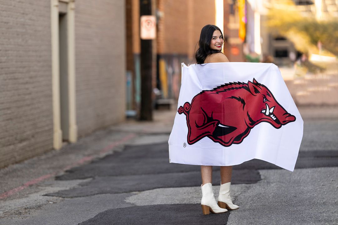 Person in black dress holds Arkansas Razorbacks flag in urban setting.