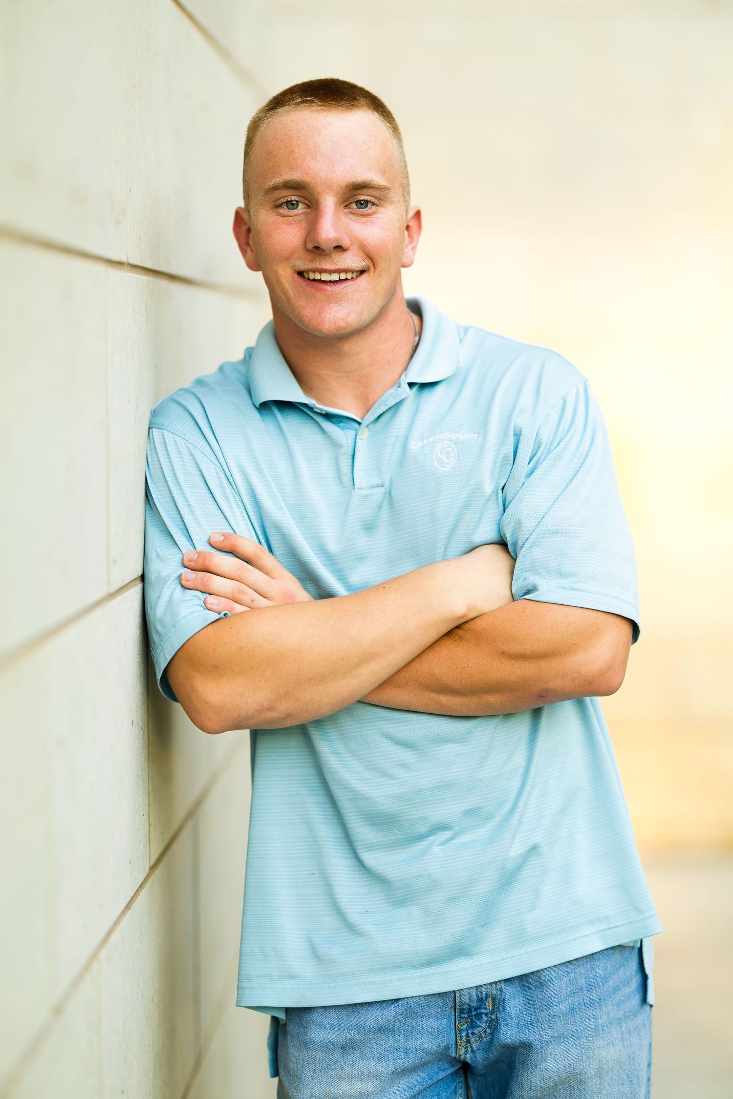 A person wearing a light blue polo shirt and jeans poses with arms crossed against a beige wall.
