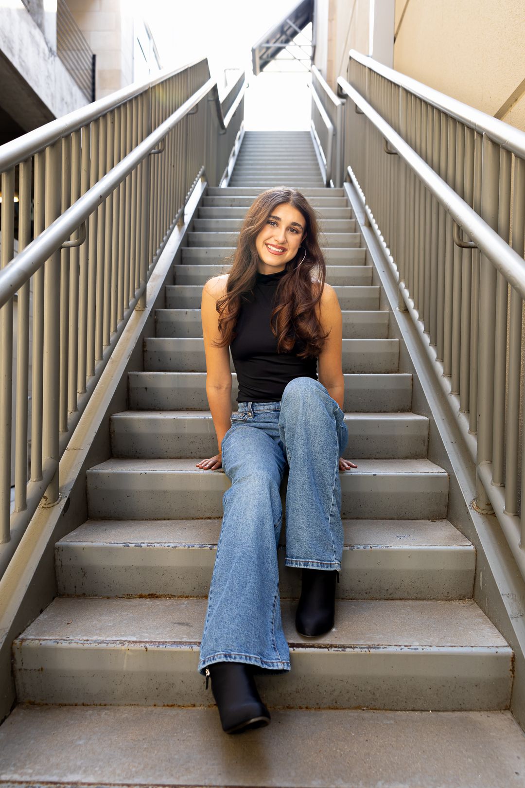 Person in casual attire sitting on concrete stairs with metal railings wearing flared jeans and black top.