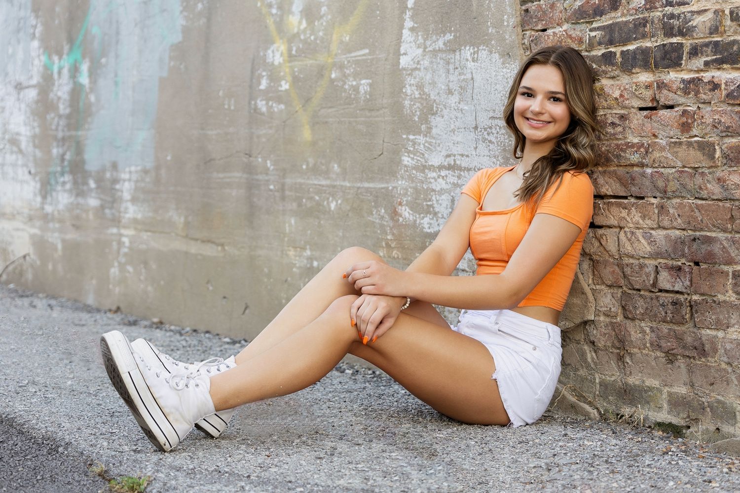 Senior portrait session featuring woman in orange top and white skirt against rustic brick wall background.