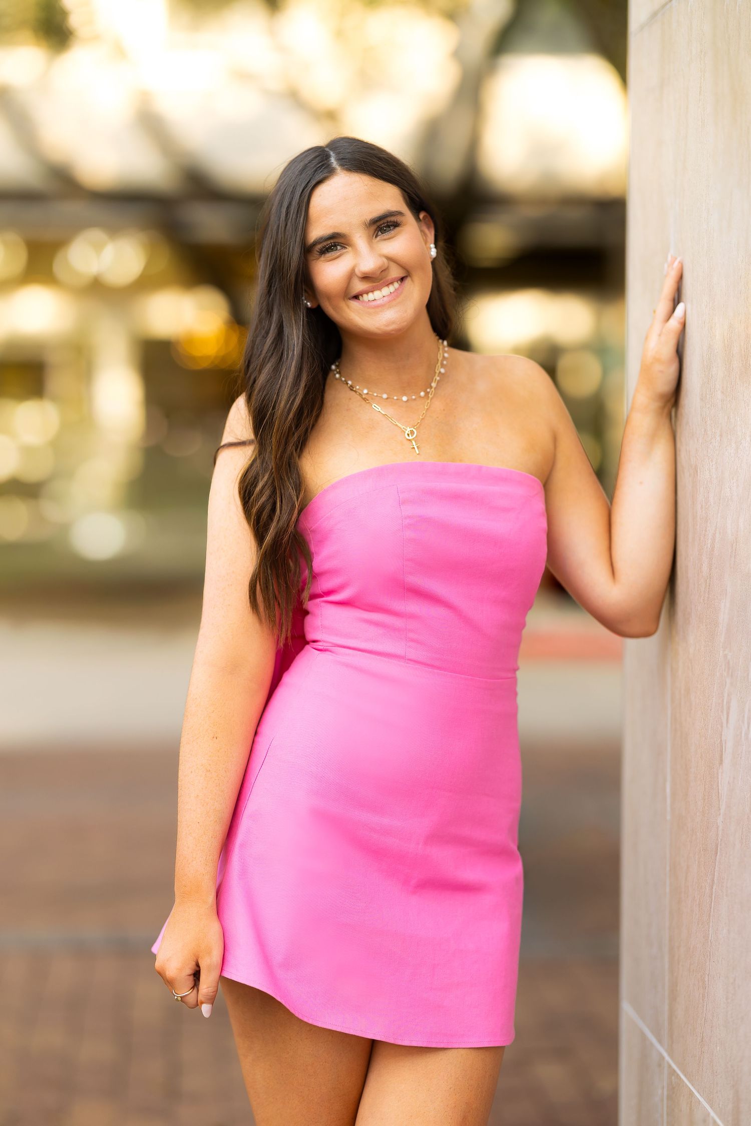 Person wearing a bright pink strapless top poses against a blurred outdoor background with warm lighting.