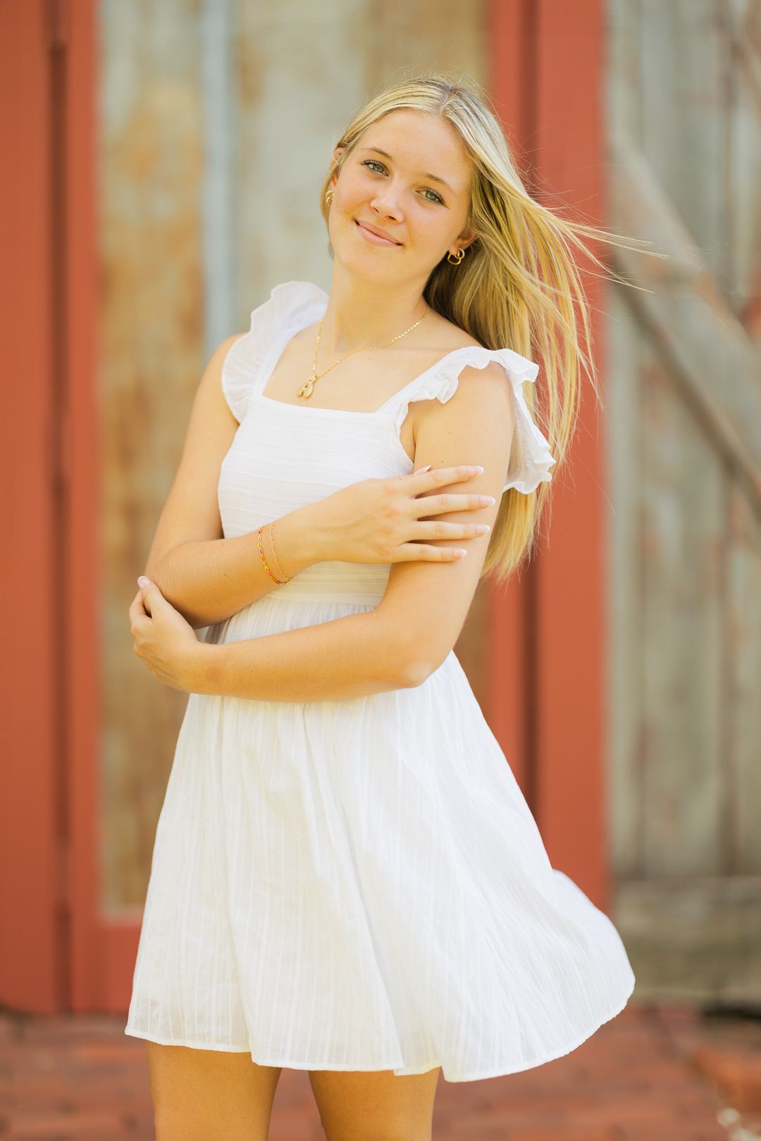 A person in a white summer dress with ruffled sleeves stands against a rustic red wall with windswept blonde hair.