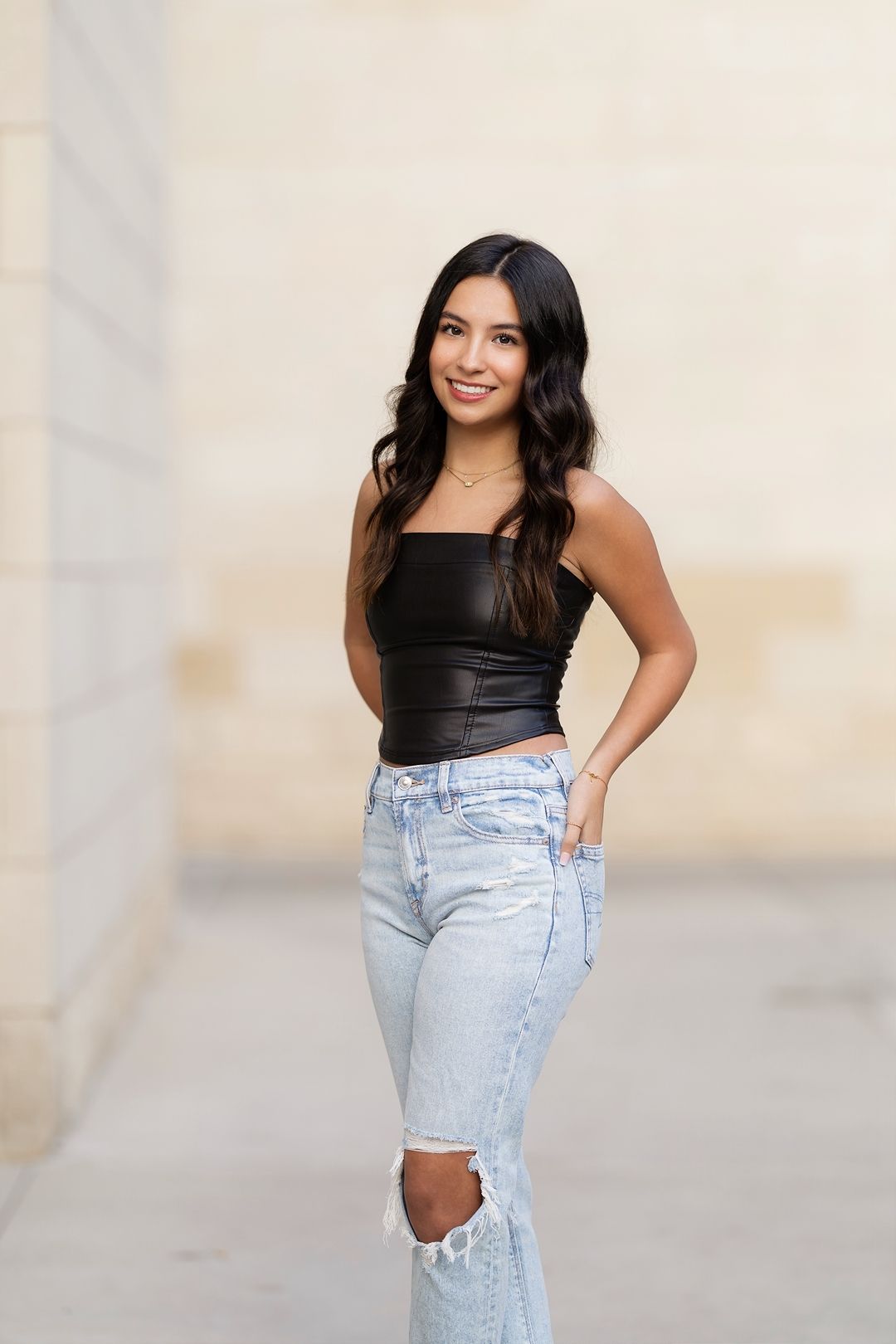 Woman in black tank top and light wash jeans poses confidently against neutral wall in casual fashion shoot.