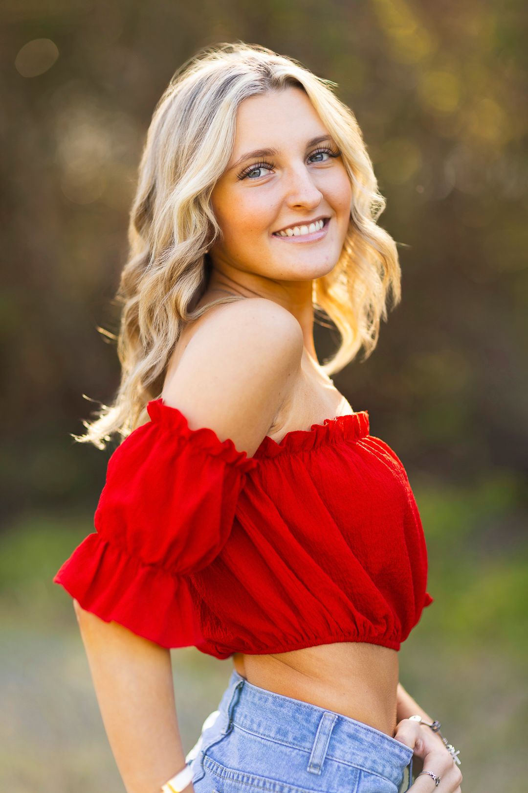 Woman wearing red off-shoulder top and jeans poses outdoors with natural bokeh background.