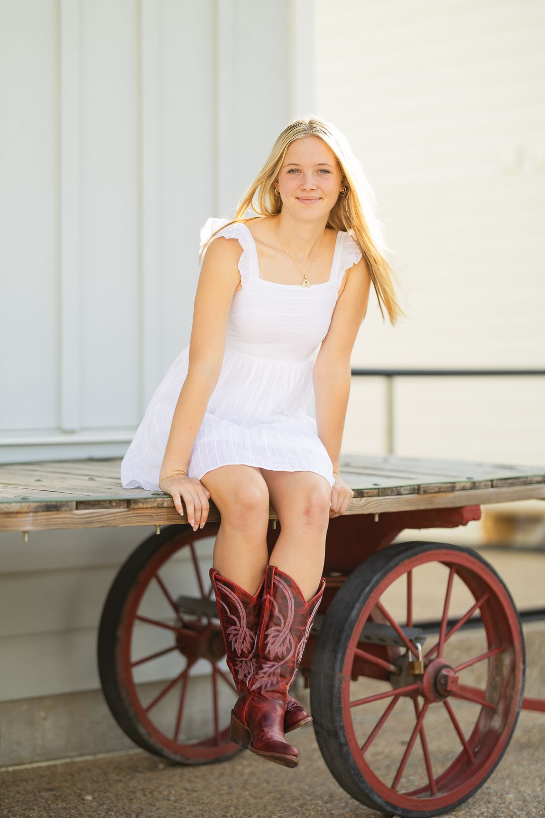 A person in a white dress and brown cowboy boots sits on a rustic wagon wheel against a white wall.