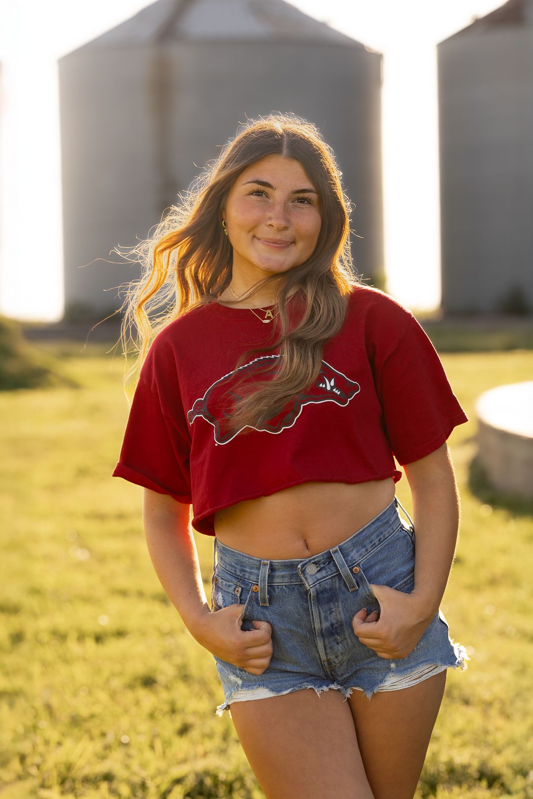 Person in red t-shirt and denim shorts poses outdoors during golden hour with sunlit background.