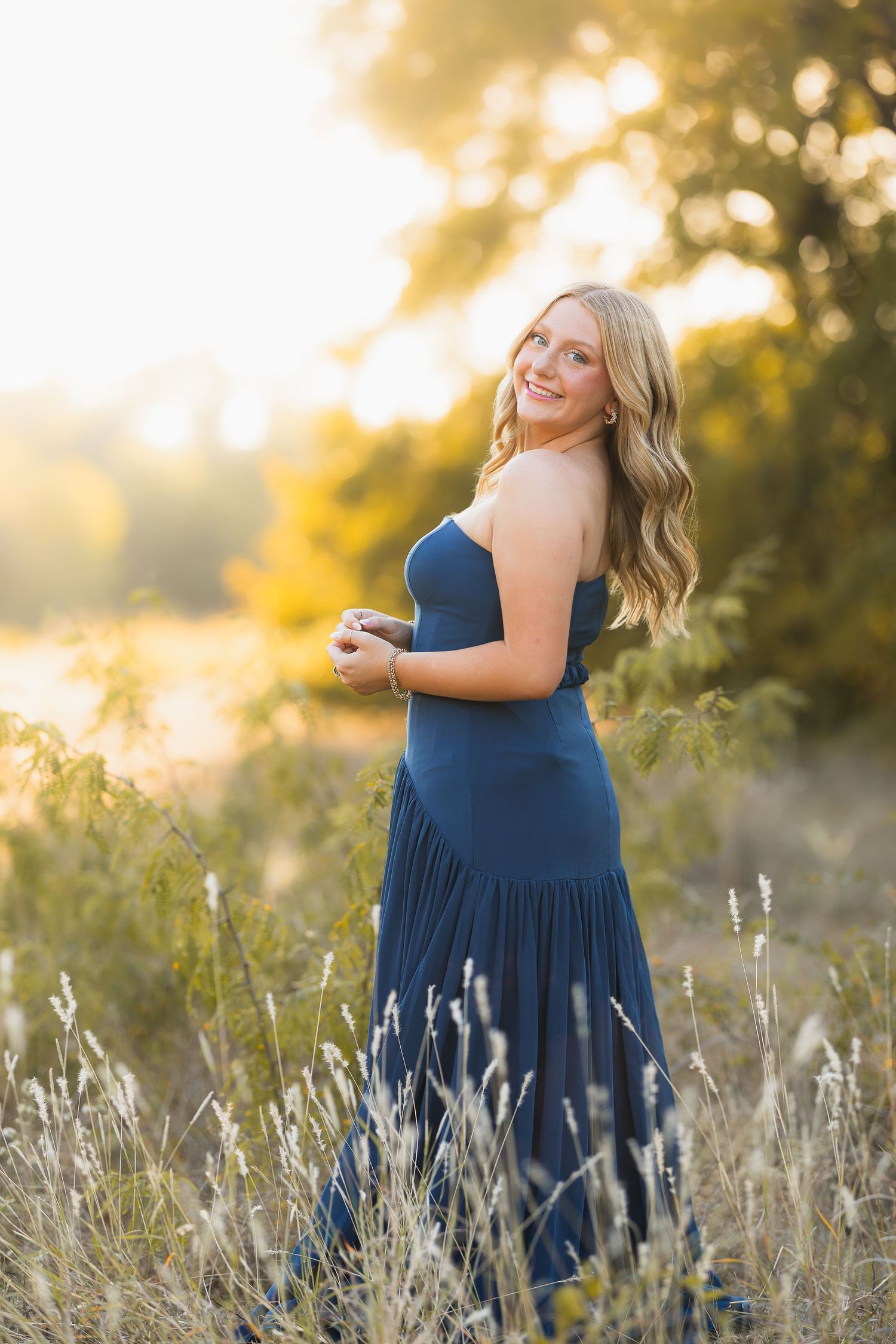 A person in a navy blue dress poses joyfully in a sunlit field of tall grass during golden hour.