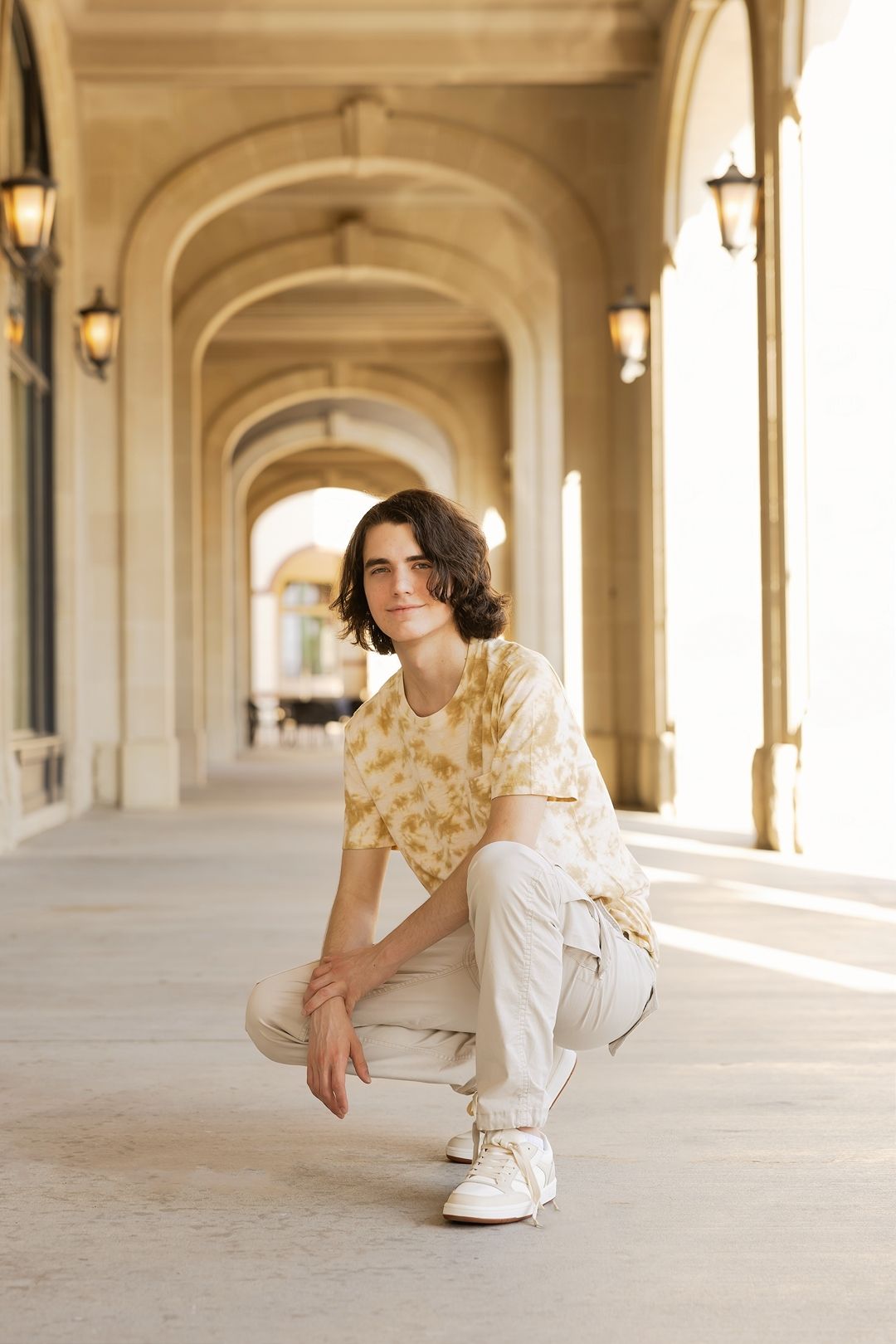 A person in white clothing and gold accents poses in an elegant arched hallway with warm lighting.