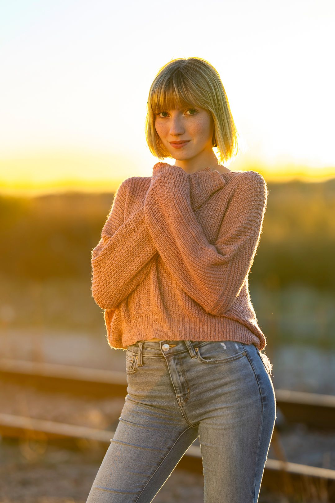 Figure in pink sweater and jeans poses against sunset backdrop.