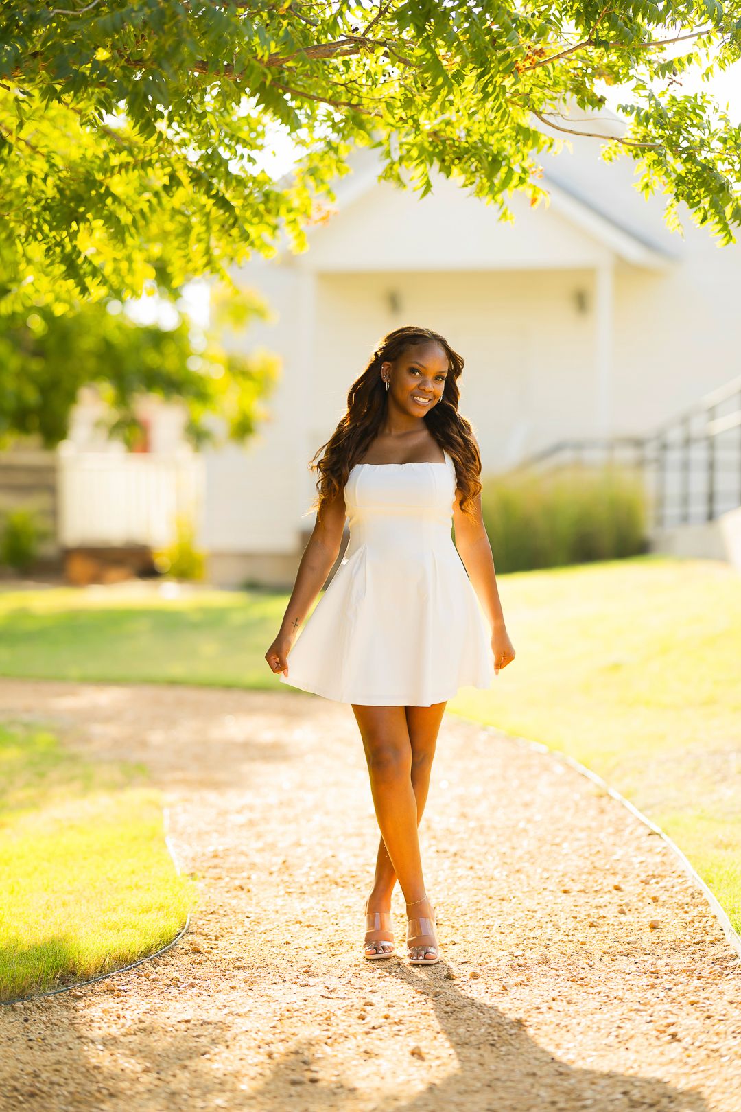 A person in white walks along a sunlit path beneath green tree branches.