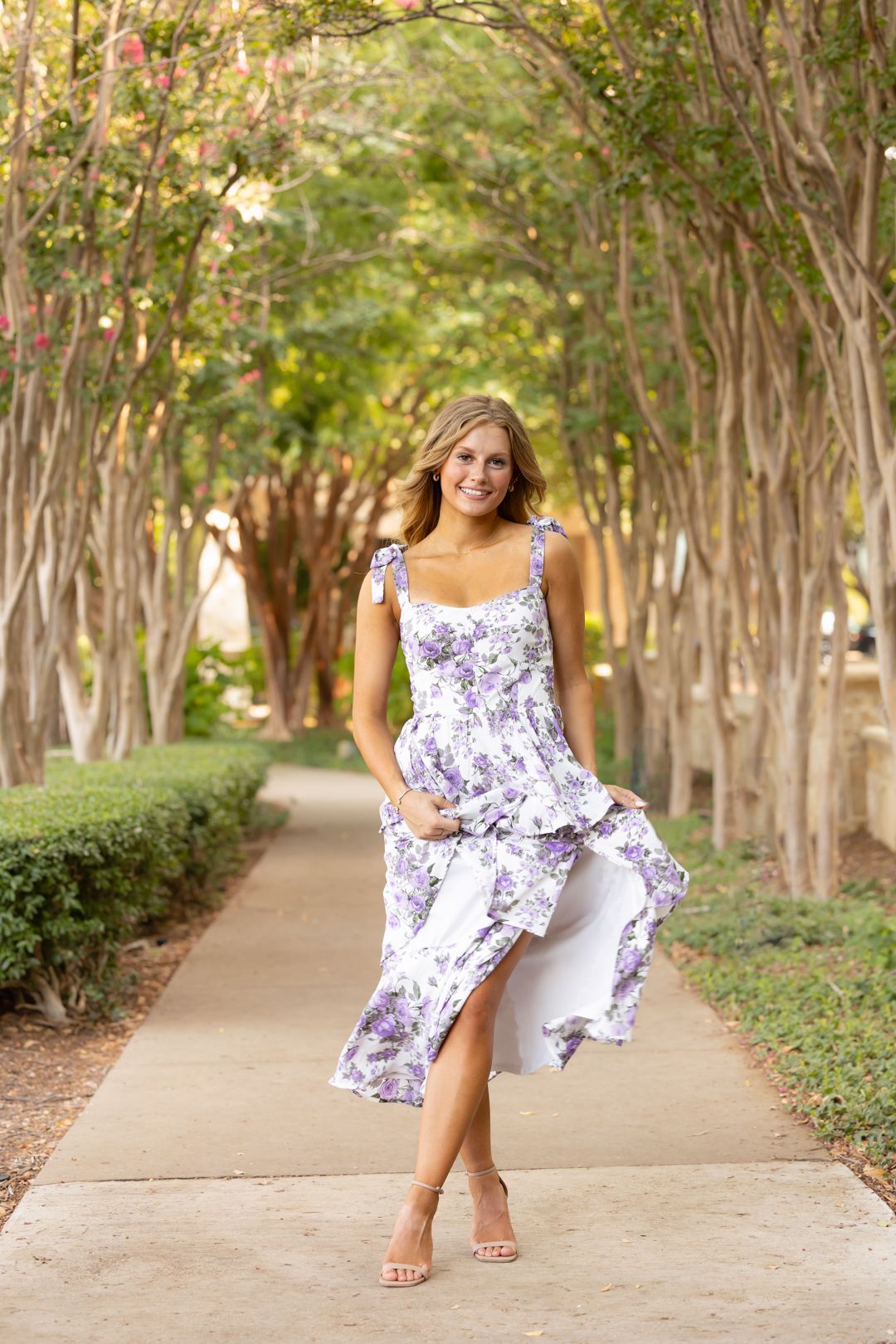 A woman in a floral dress poses playfully in a series of photos along a tree-lined pathway on a sunny day.