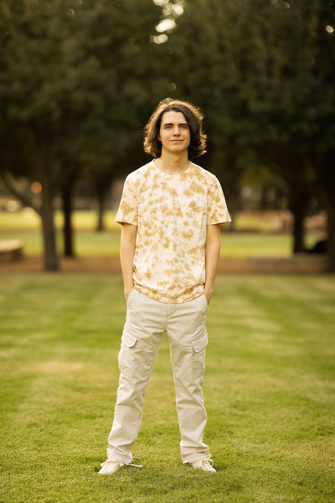 Person in tie-dye shirt and white cargo pants stands on grass in a park with trees in background at sunset.