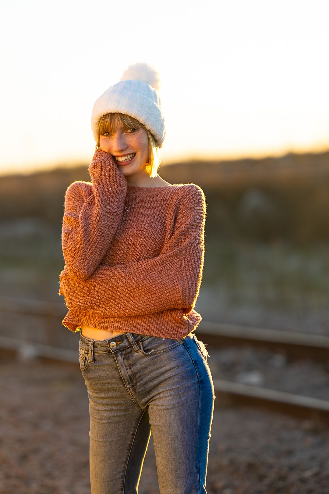 Person wearing orange sweater and jeans poses outdoors during golden hour sunset with winter beanie hat.