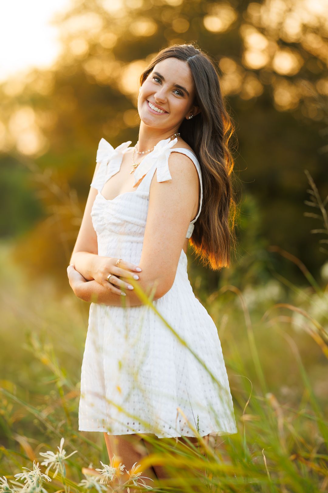 Young woman in white dress smiling outdoors in golden hour sunlight surrounded by wildflowers and green trees.