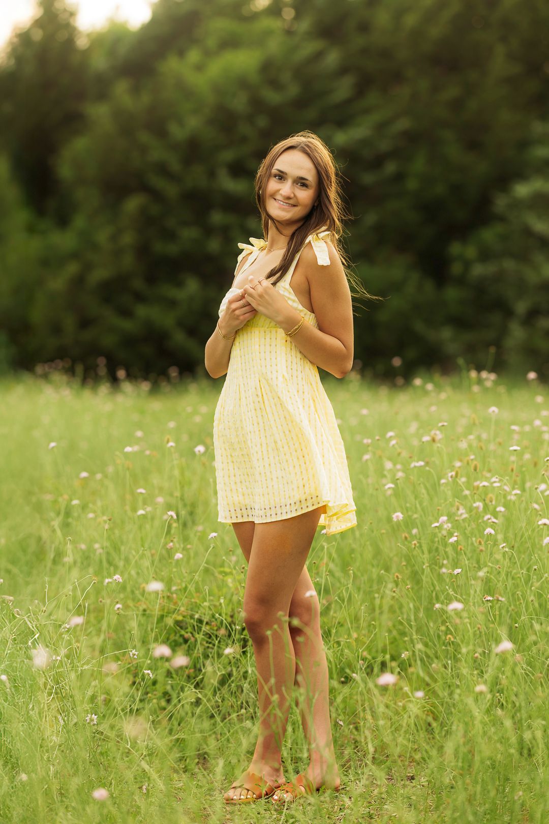 Young woman in yellow sundress standing in green meadow with wildflowers, smiling outdoors near trees.