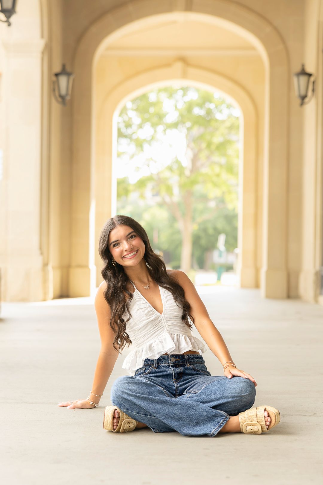 Person in white top and jeans sits casually under arched colonnade.