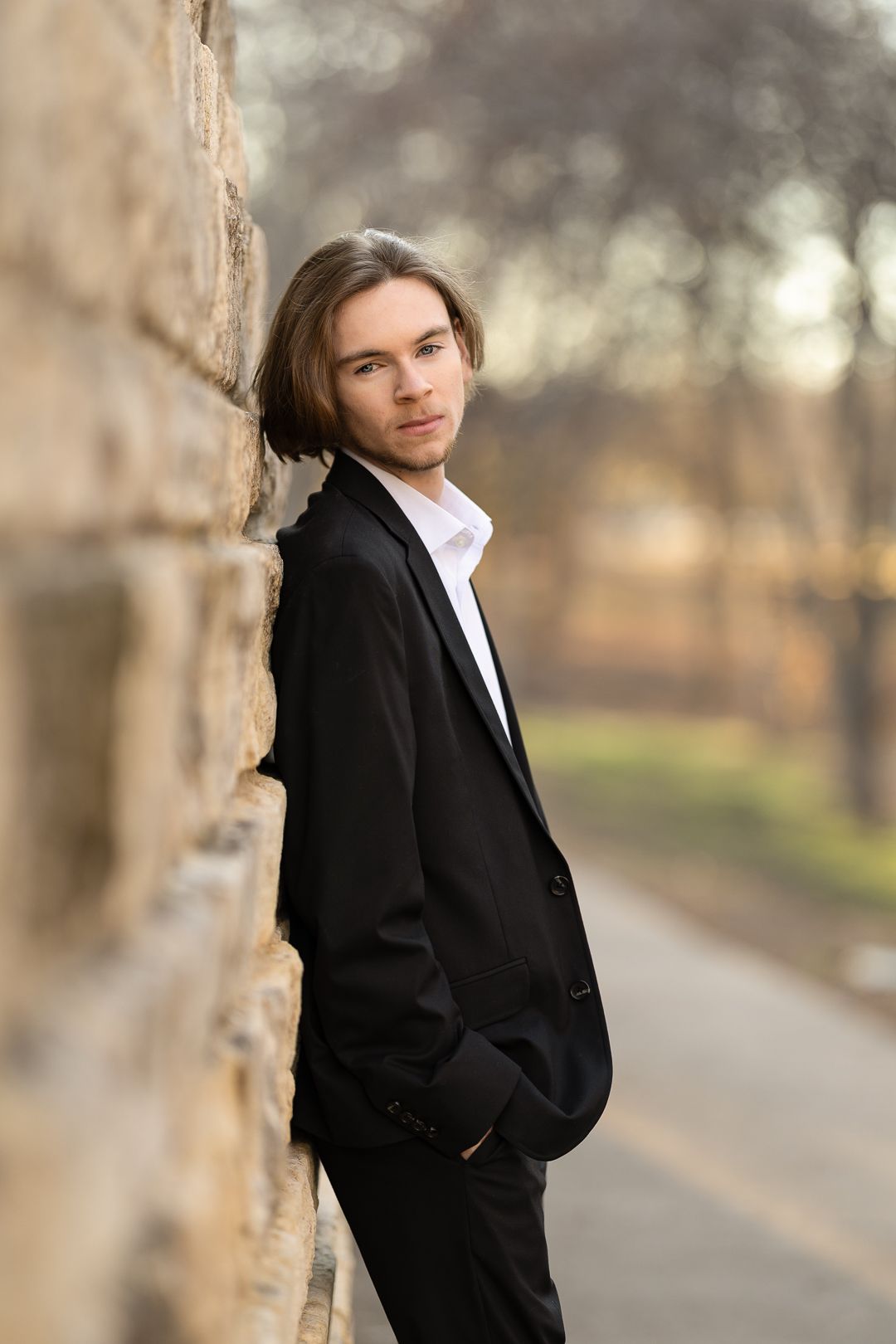 A person in a black suit jacket leans against a tree trunk along a misty park pathway.