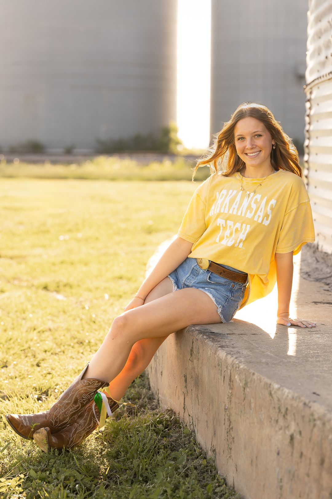 Young woman in yellow shirt and denim shorts relaxes on concrete ledge during golden hour sunset.