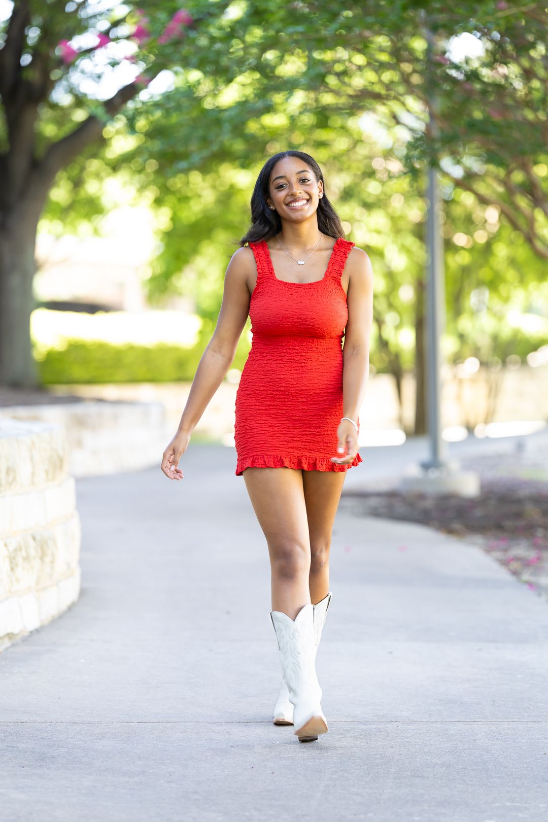 A person in a bright red top and white boots poses playfully on a tree-lined sidewalk on a sunny day.