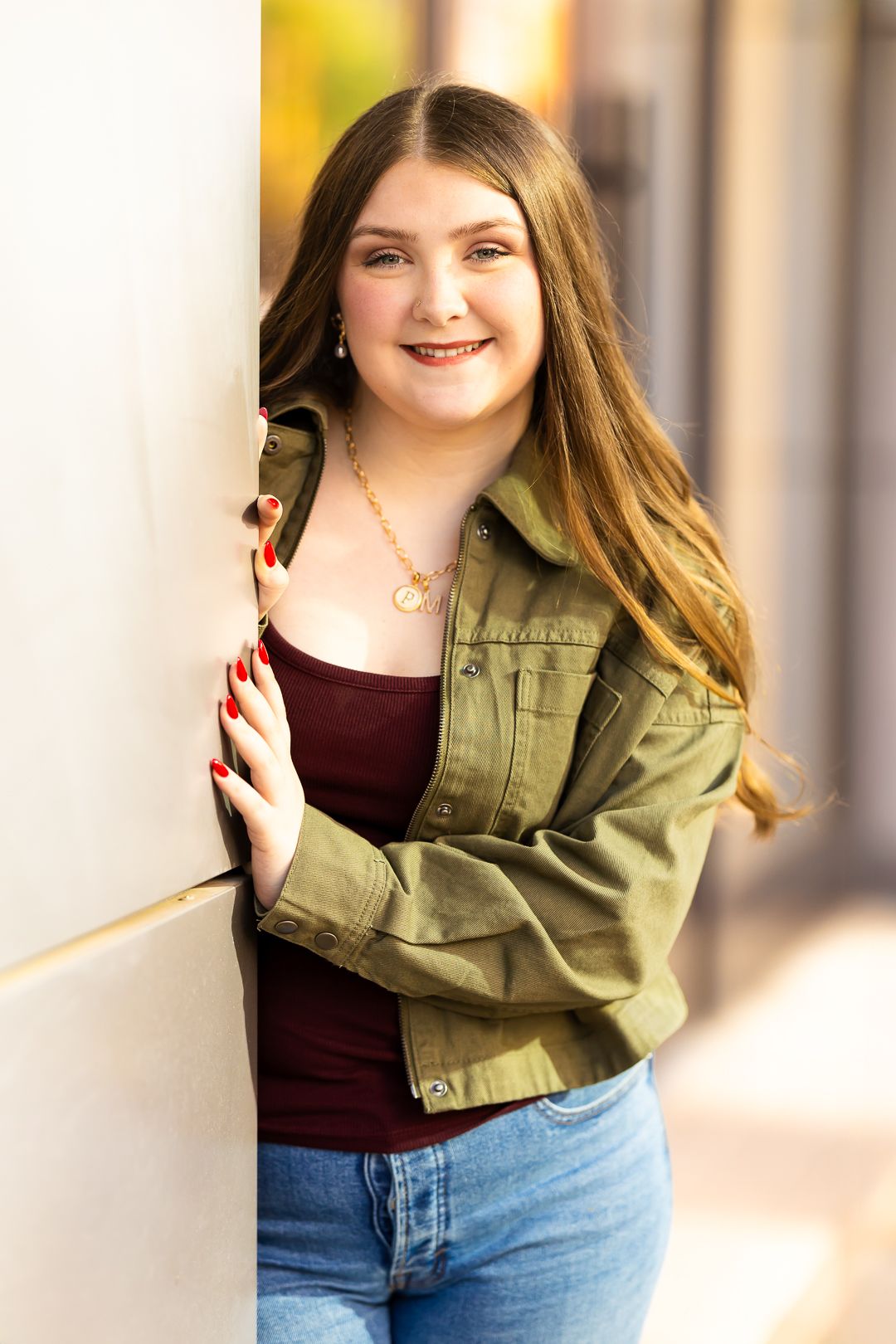 A person wearing an olive green jacket and burgundy top poses against a wall in natural lighting.