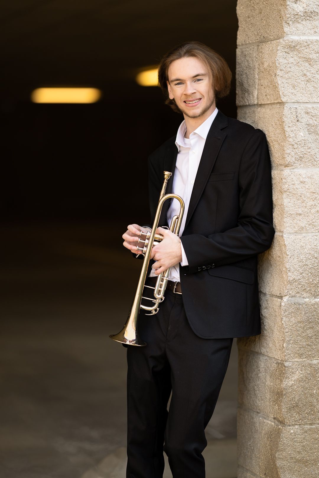 Professional musician in formal black attire poses with trumpet against stone wall in artistic portrait series.