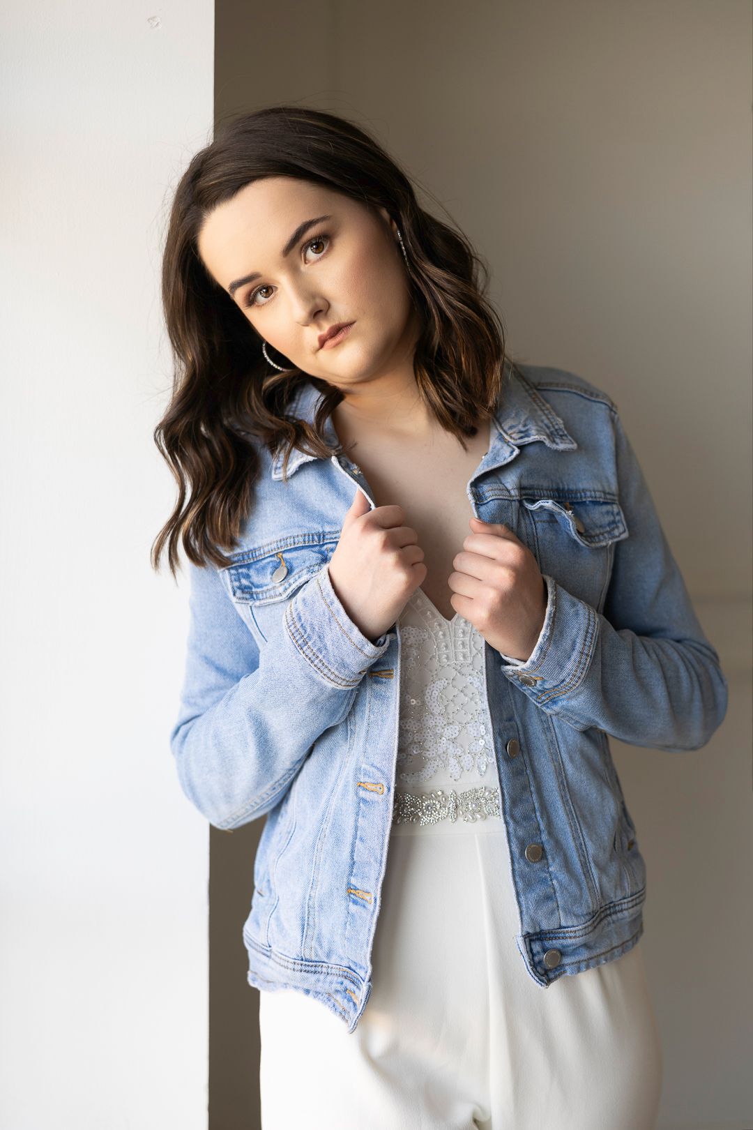 Person wearing denim jacket and white dress poses against neutral background.