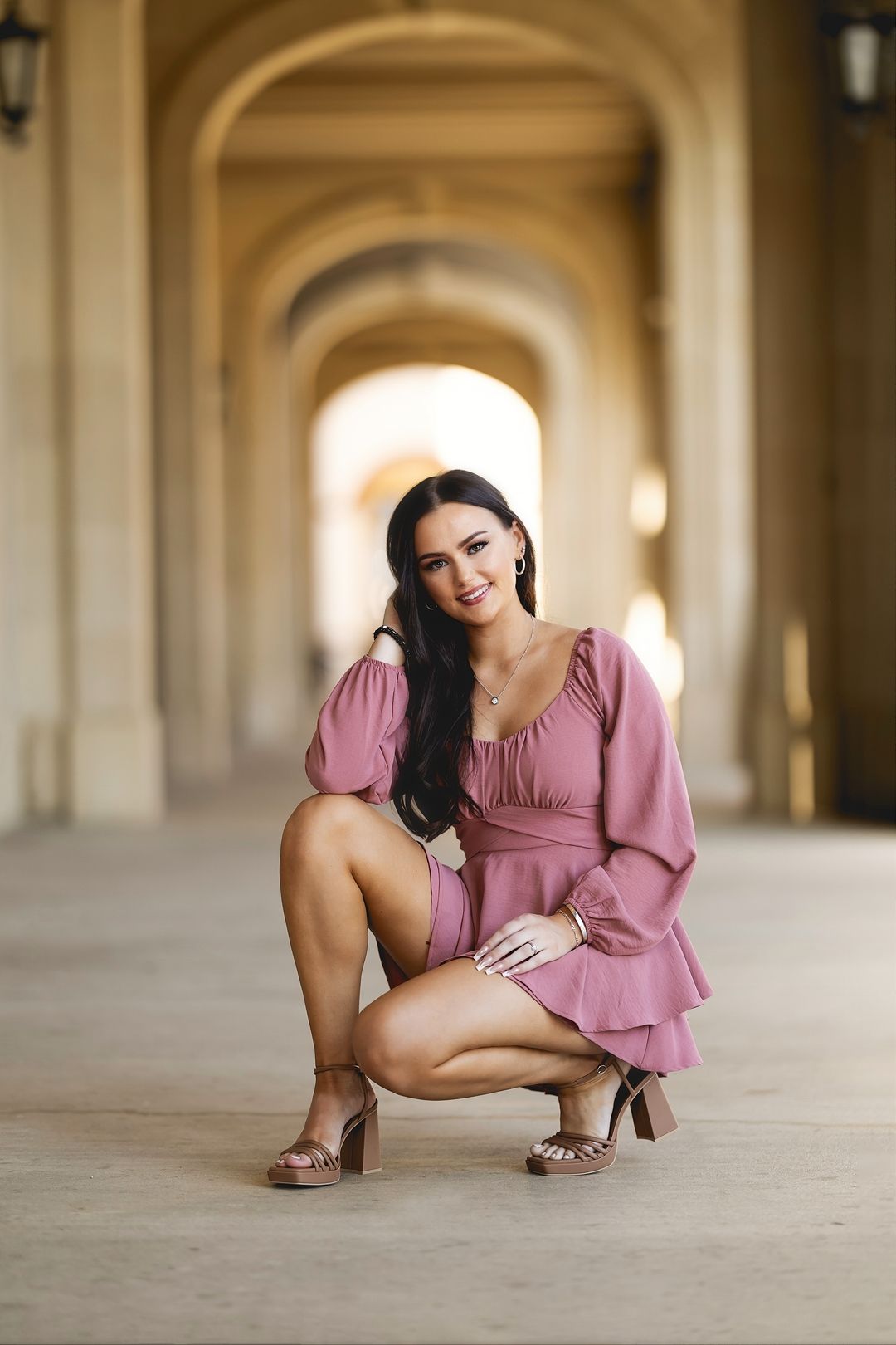 A woman in a pink dress poses gracefully on the floor in an elegant hallway with arched architecture.