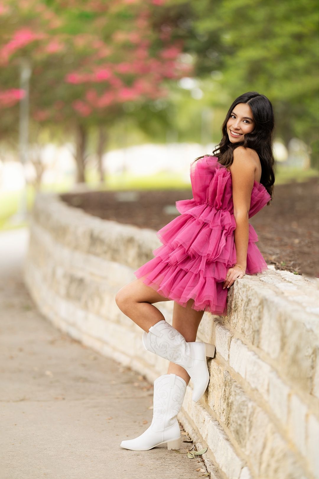 A person in a pink ruffled dress and white boots poses on a stone wall in an outdoor garden setting.