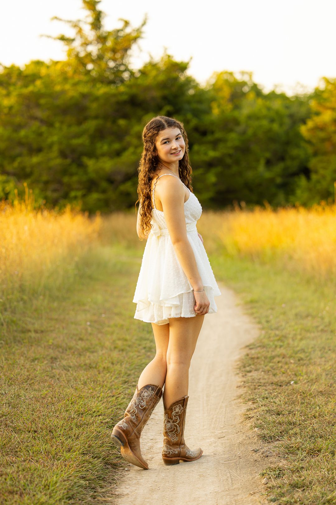 A child in a white dress and brown cowboy boots stands on a dirt path during golden hour.