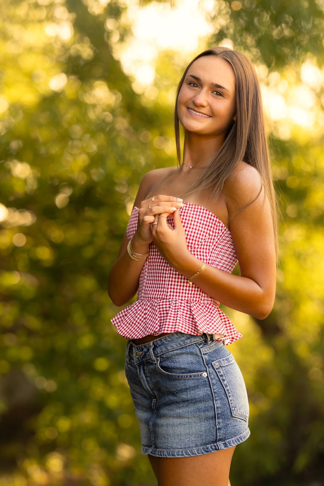 Person in red gingham top and denim shorts poses outdoors during golden hour with blurred natural background.