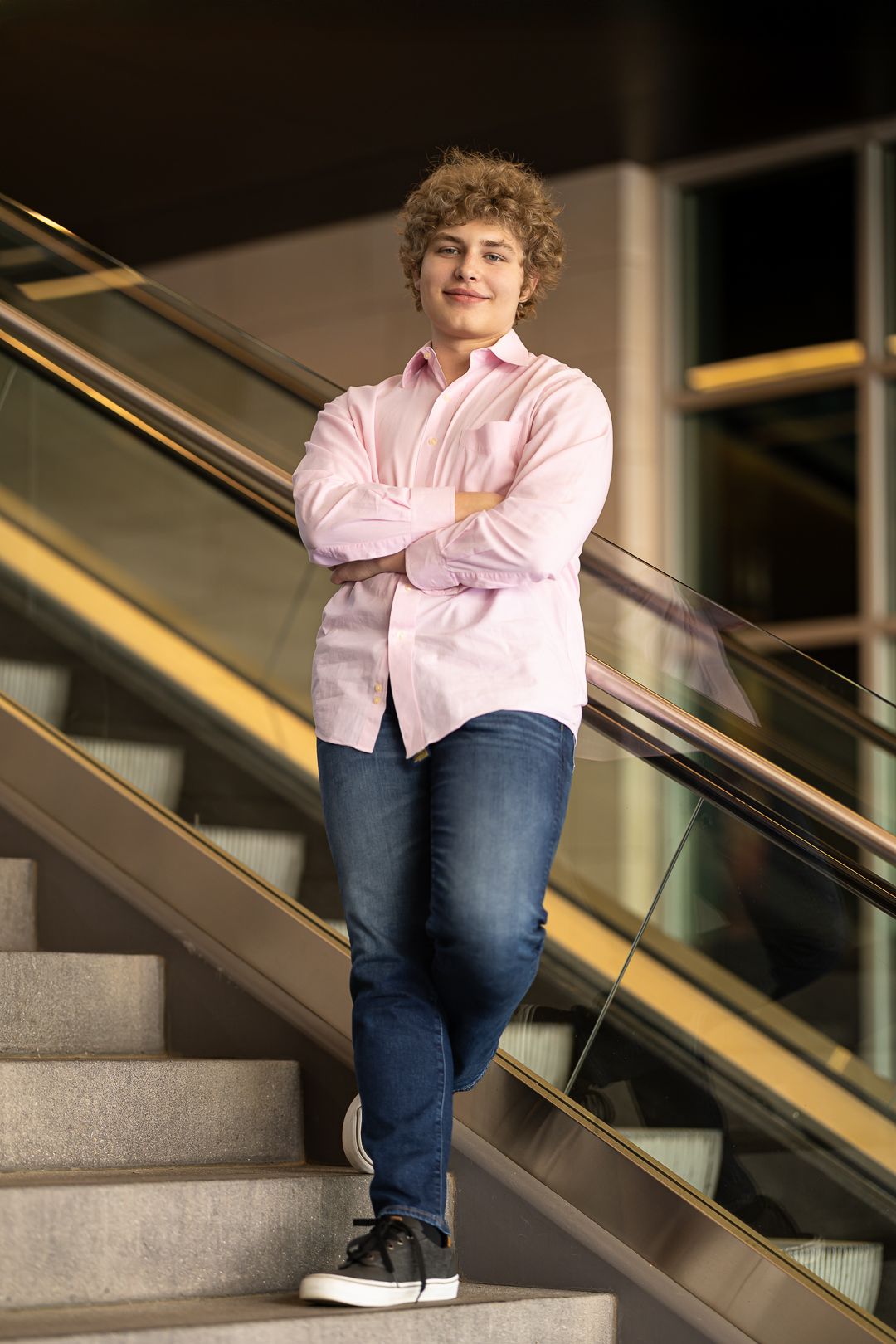 A person in a pink button-down shirt and jeans stands casually on stairs with arms crossed, leaning against an escalator railing.