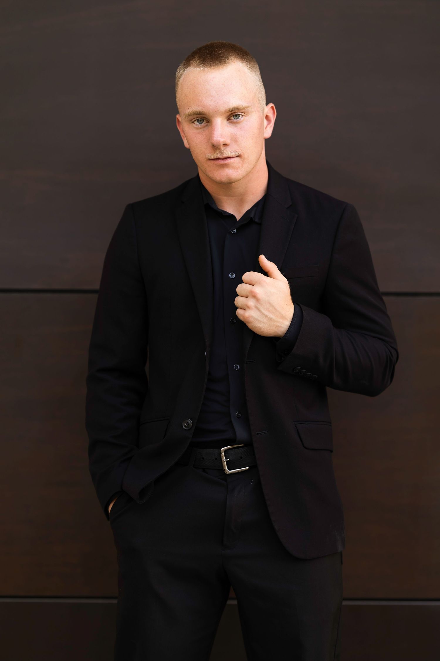 A person wearing a black suit jacket poses against a dark background in a professional headshot series.