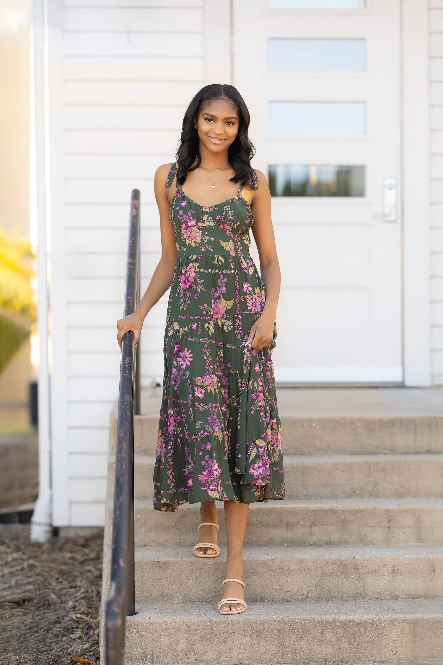A floral sundress in olive green with pink flowers is styled with beige sandals on concrete steps.