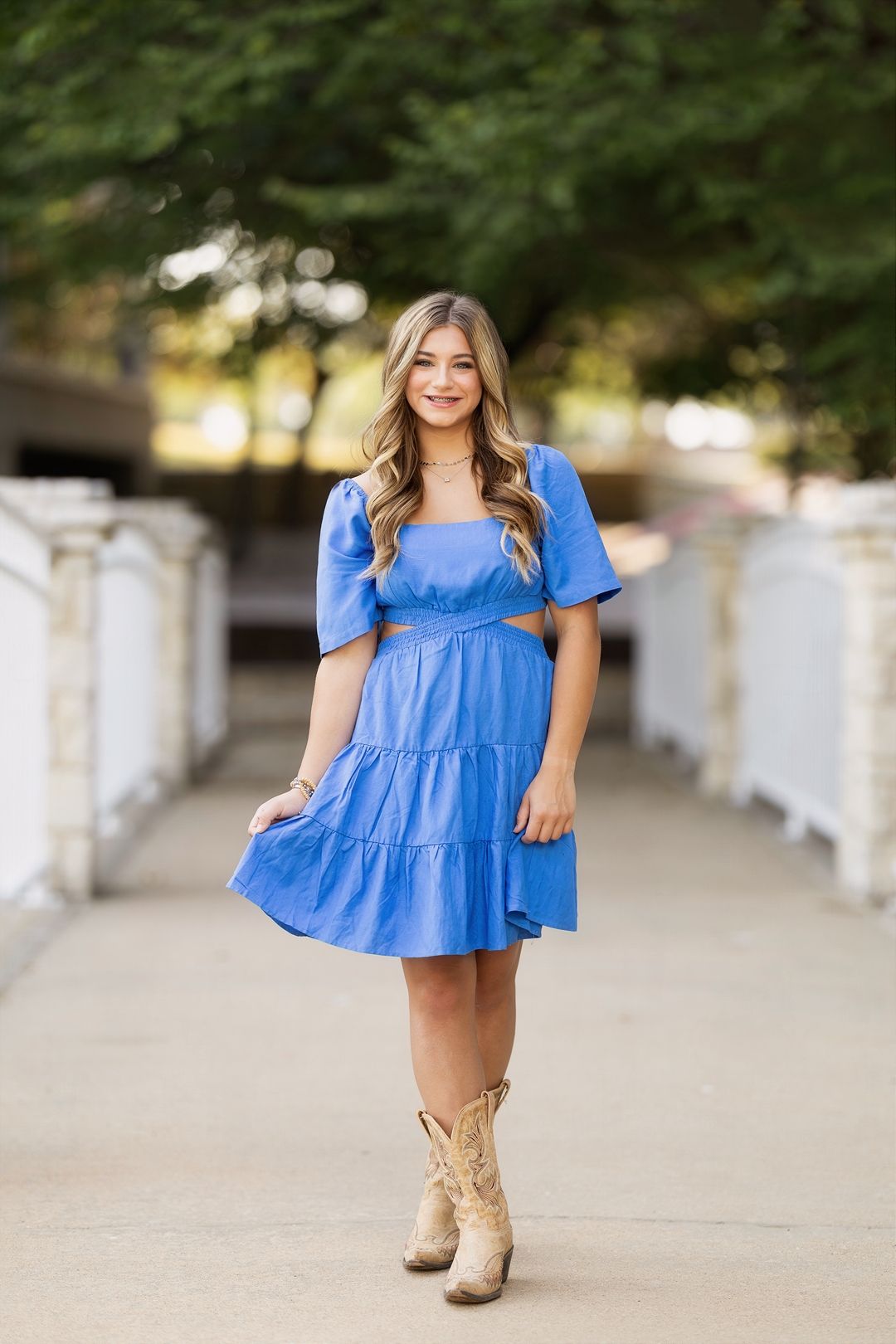 A person wearing a bright blue tiered babydoll dress and tan boots poses outdoors against a blurred background.
