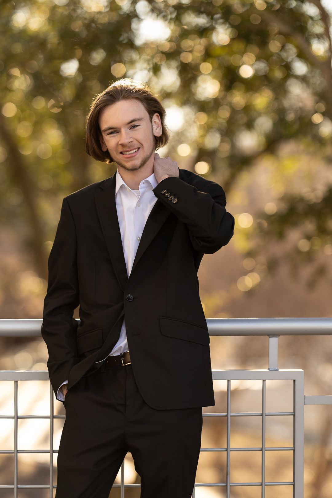 A person in a black suit leans against a railing outdoors with a blurred bokeh background.