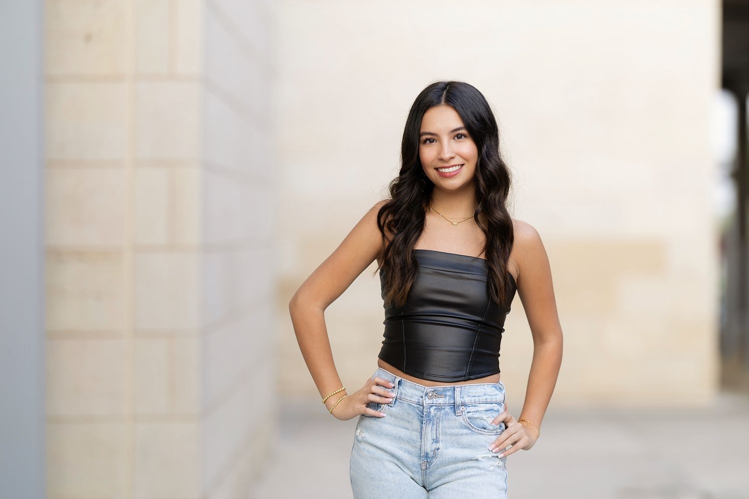 A person in a black tank top and light jeans poses against a light colored wall with a bright smile.