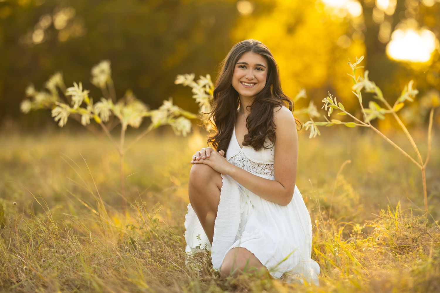 A white sundress-clad figure sits in golden sunlit grass during a dreamy outdoor portrait session at sunset.
