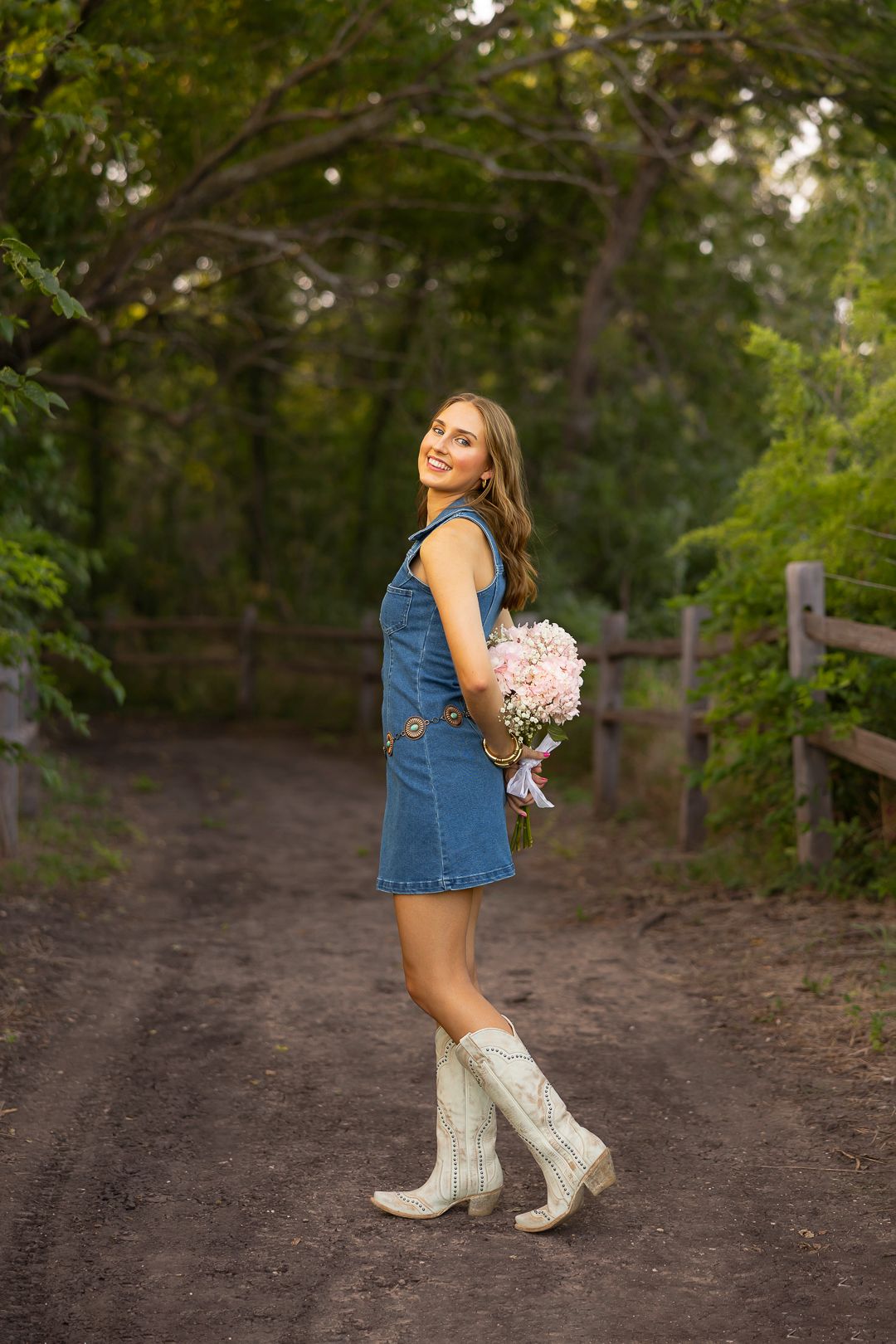 Outdoor portraits in denim dress and white boots holding pink flowers on a wooded path.