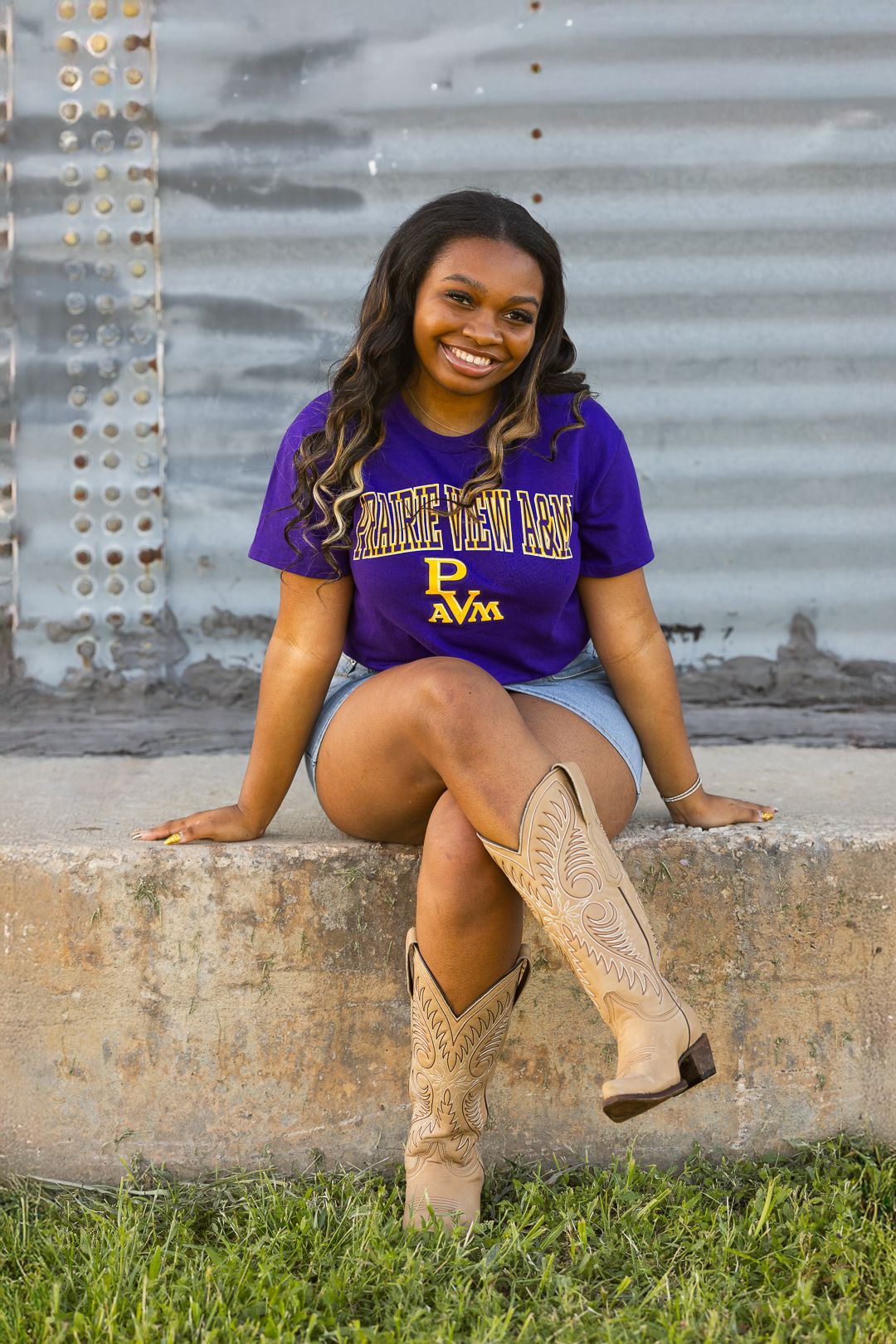Student in purple university shirt and tan boots sits casually on concrete ledge near grass.