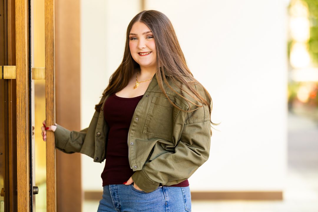A person wearing a green military style jacket and jeans poses casually in an outdoor setting with warm lighting.