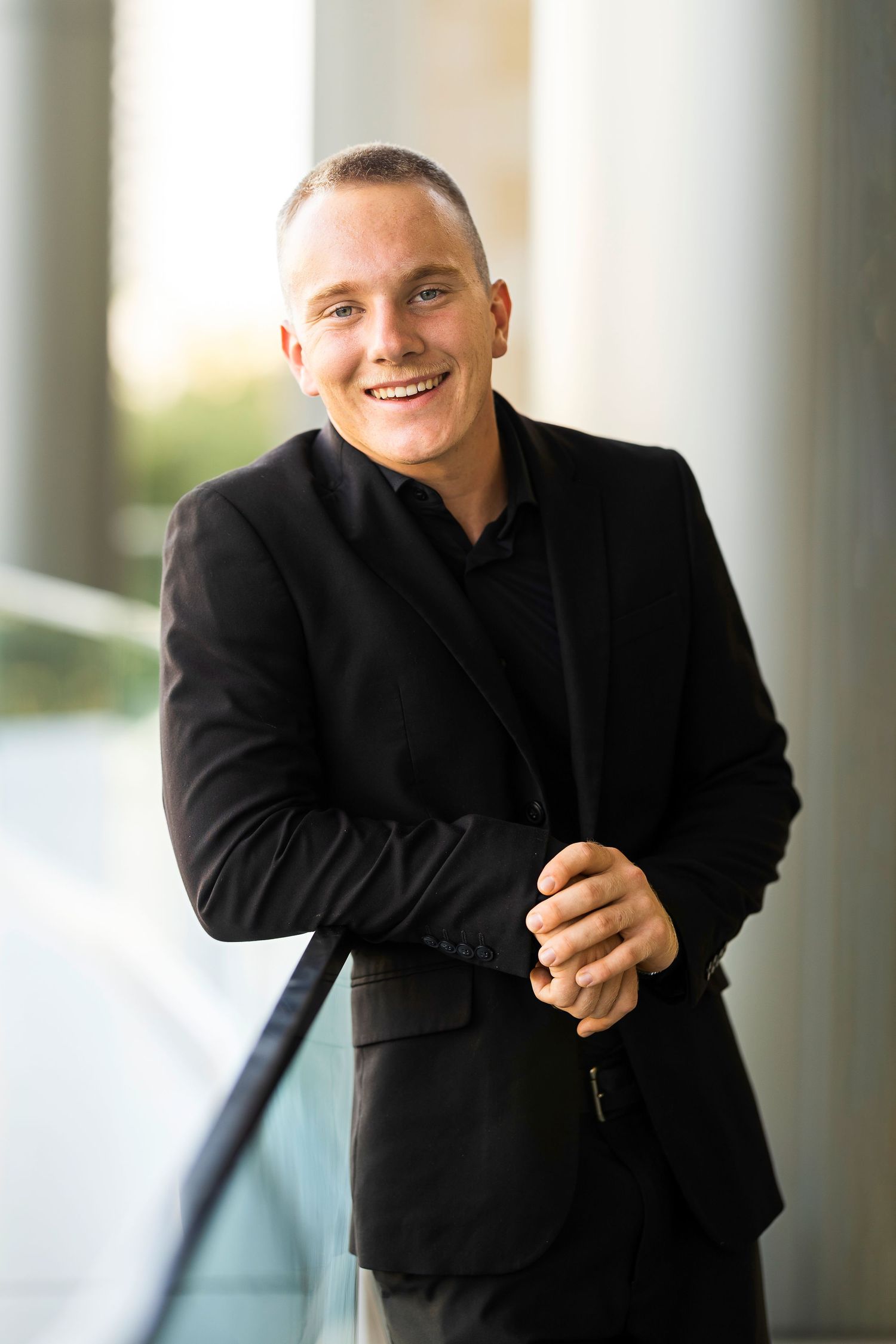 A person in a black suit jacket smiles warmly while leaning against a bright architectural column.