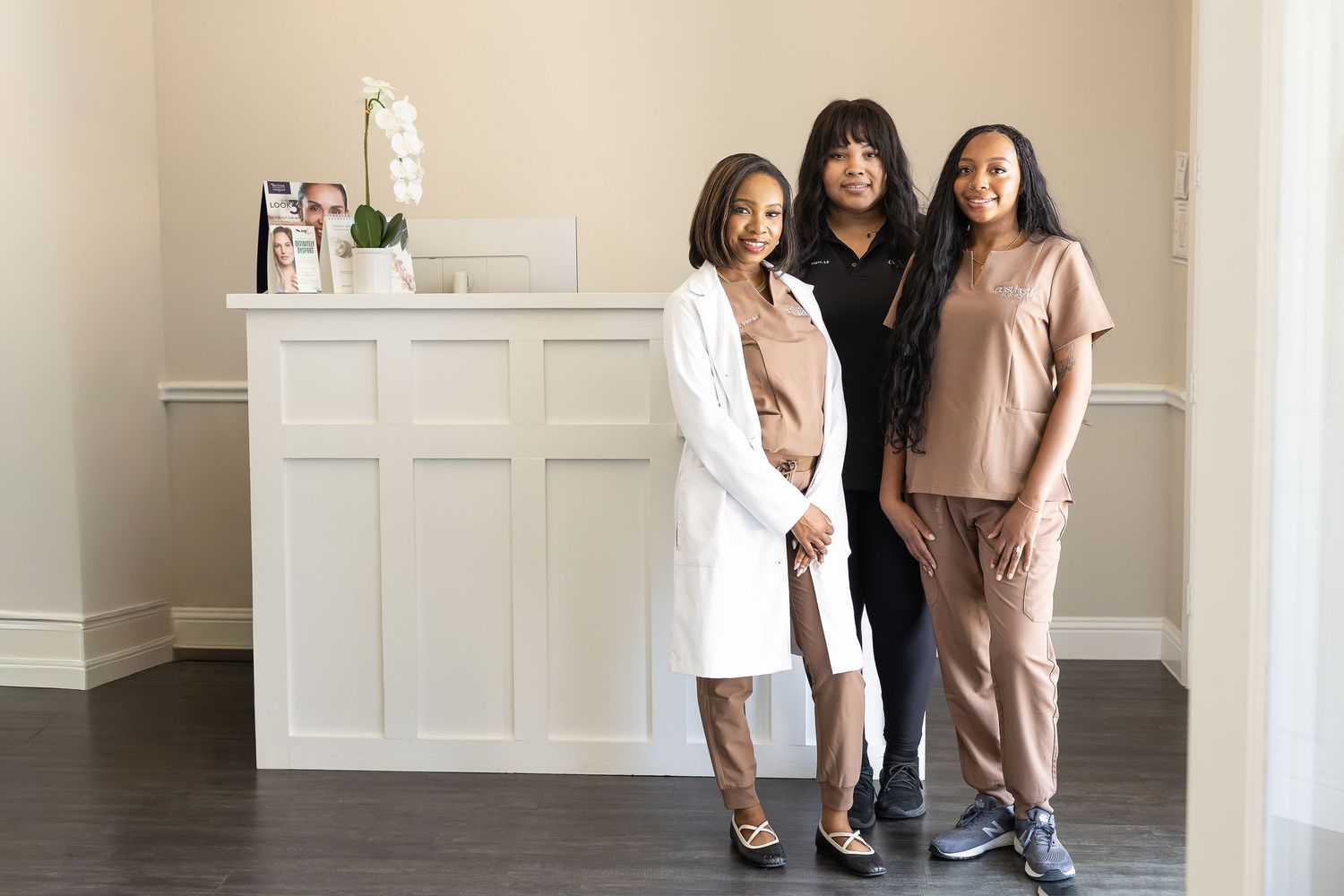 Three people in coordinating neutral outfits pose together in a bright studio space with white walls and wood floors.