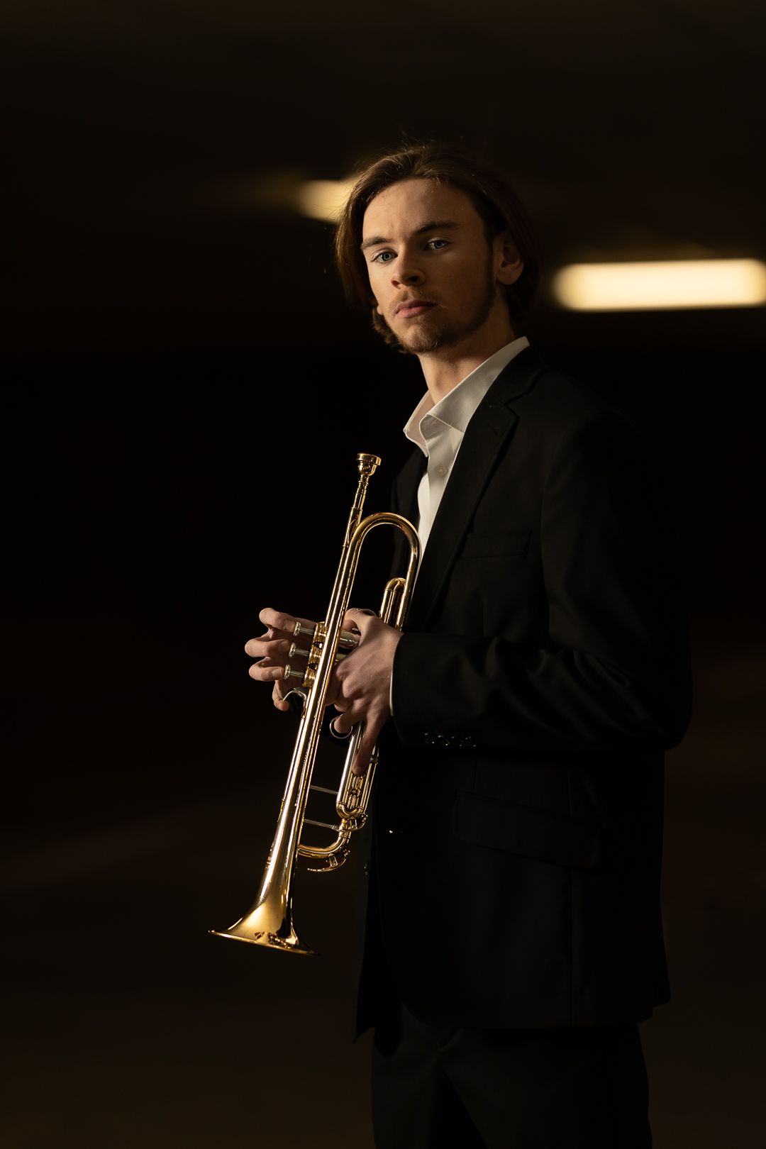 A musician in formal black attire holds a brass trumpet in dramatic low-key lighting.