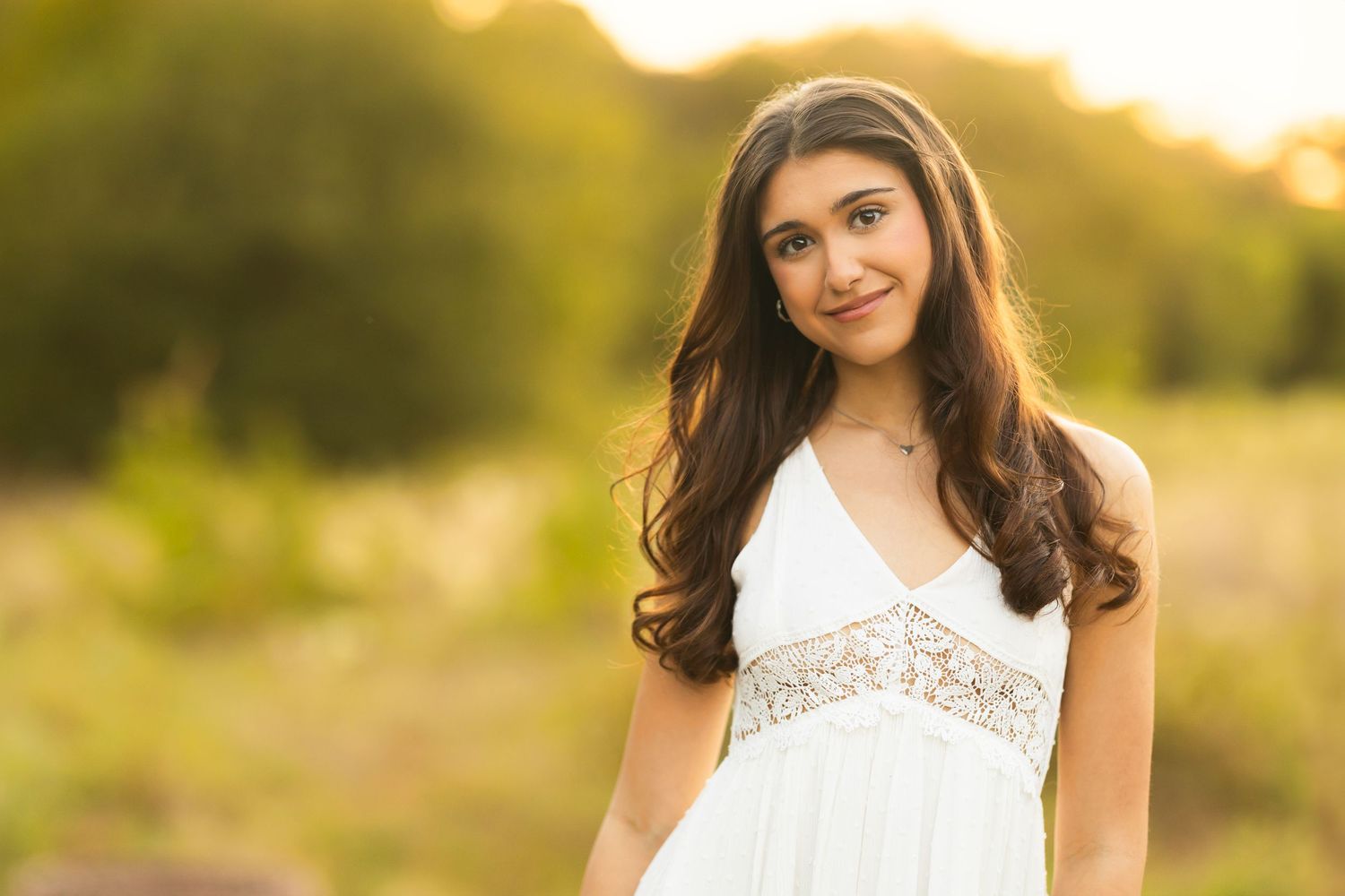 A woman in a white lace dress poses outdoors during golden hour with warm sunlight creating a dreamy atmosphere.