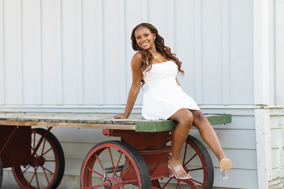 Woman in a white dress sits gracefully on vintage red wagon wheels against a light colored background.