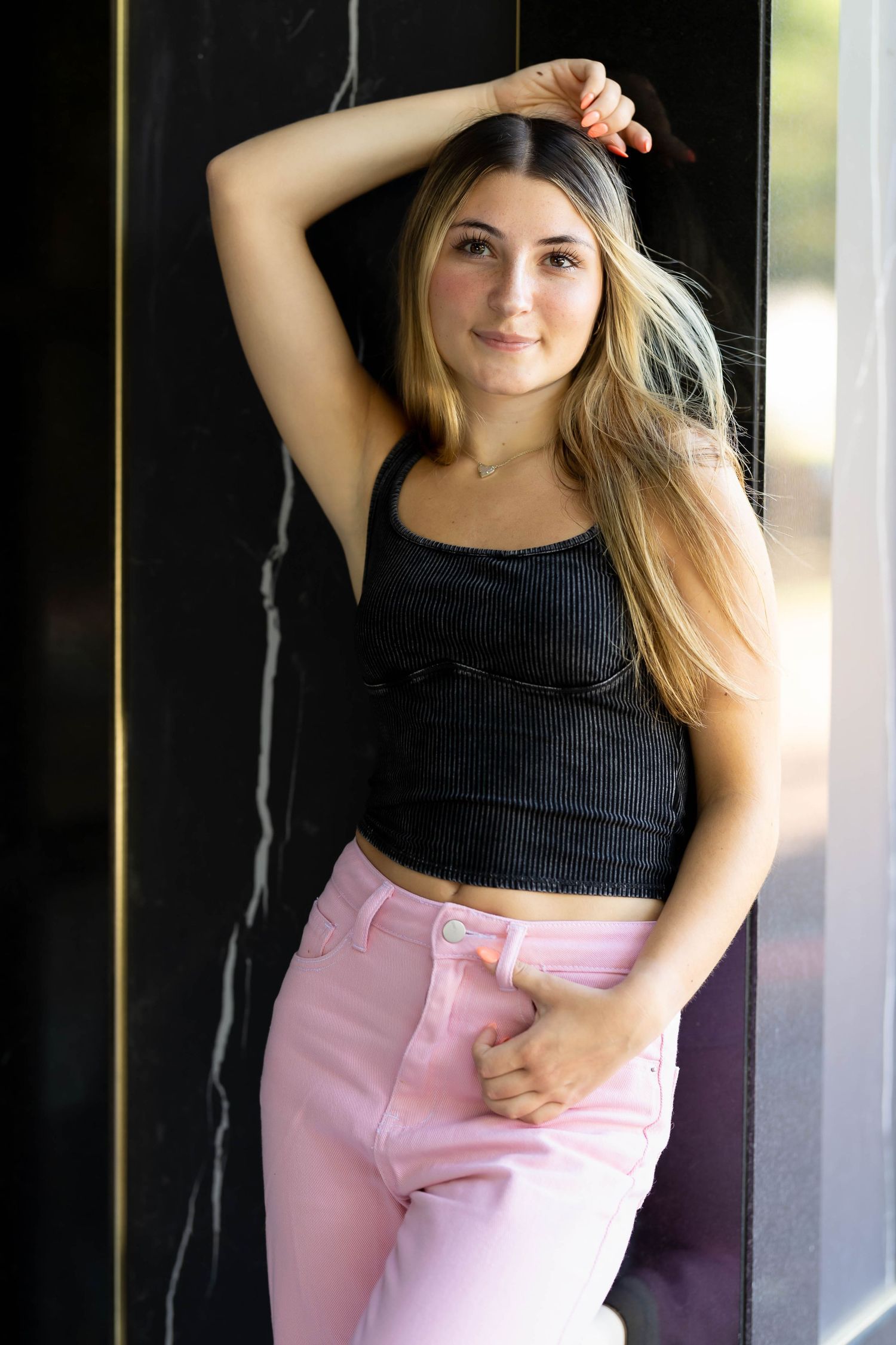 Portrait of a person in a black tank top and pink pants posing against a dark wall in natural lighting.