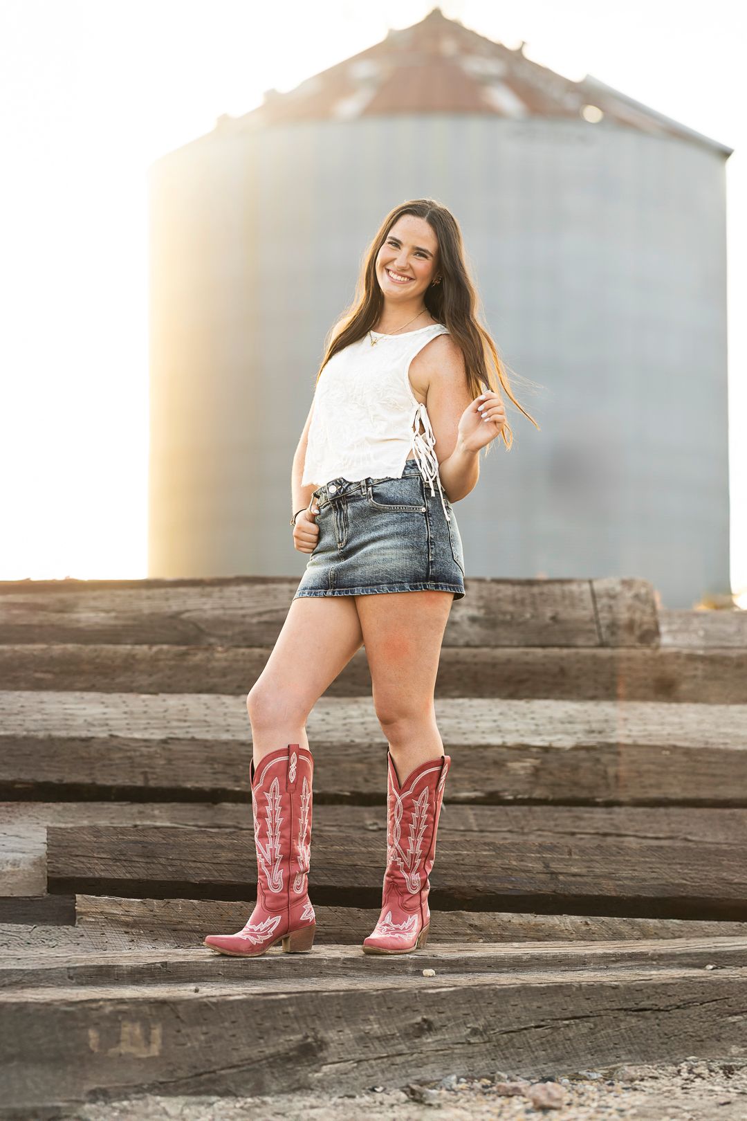 Pink cowboy boots stand out against rustic wooden steps and metal grain silos in a country setting.