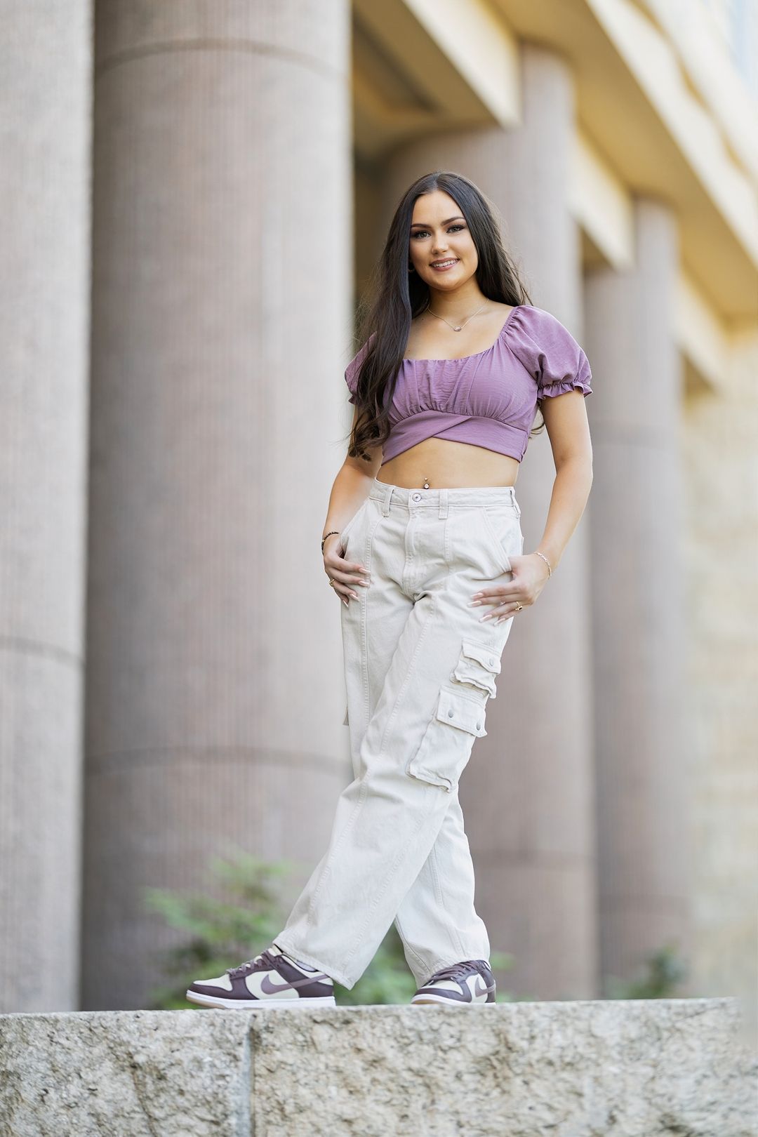 A person wearing a purple crop top and white cargo pants poses casually against stone columns in an outdoor setting.