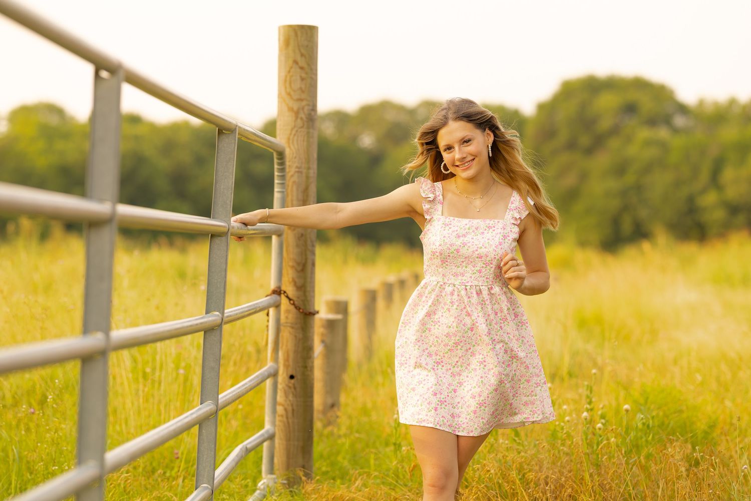 A person in a flowy pink dress runs playfully along a wooden fence in a sunlit meadow during golden hour.