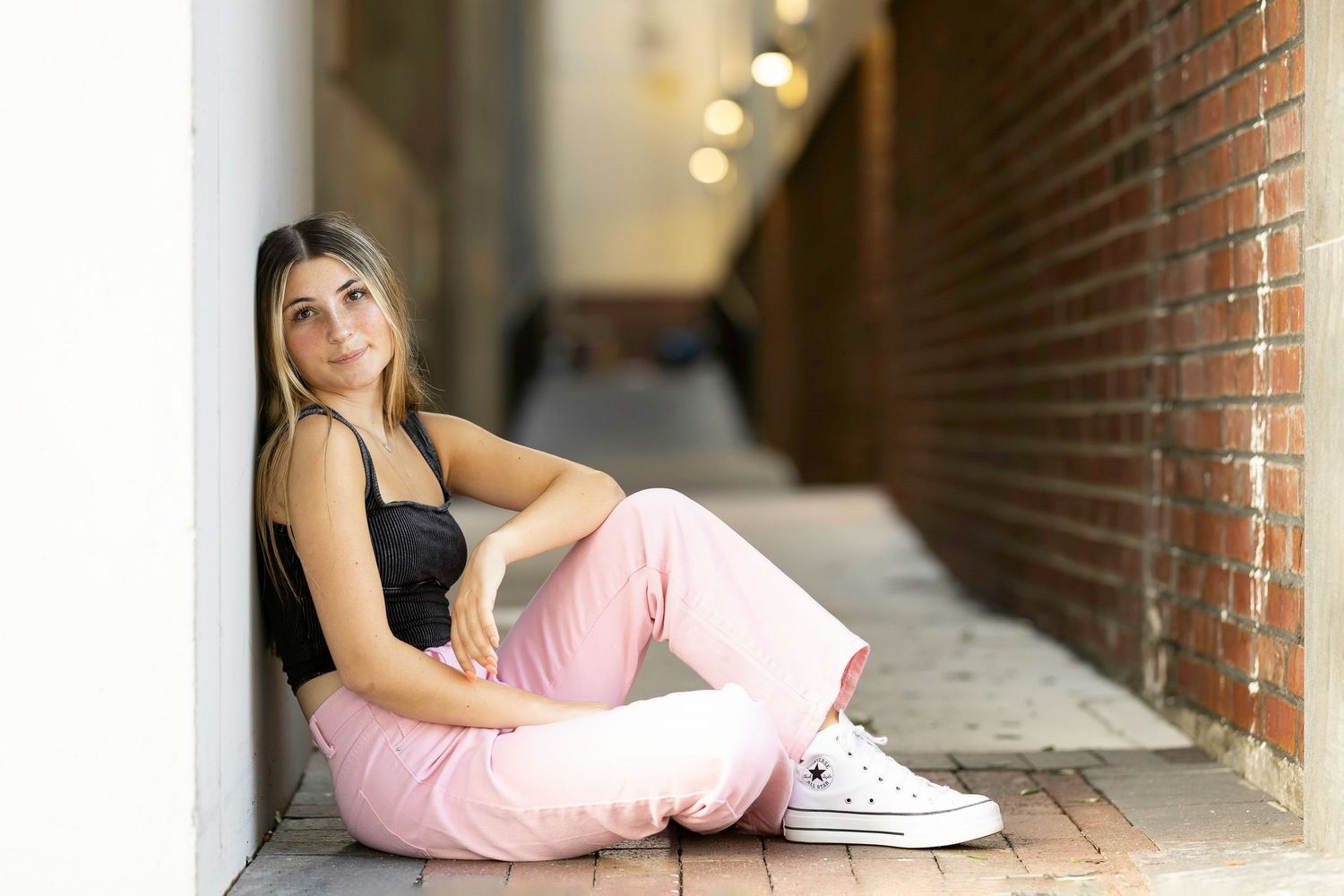A person in pink pants and black top sits against a brick wall in an urban alleyway wearing white sneakers.